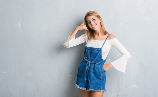 Beautiful young woman standing over grunge grey wall smiling doing phone gesture with hand and fingers like talking on the telephone. Communicating concepts.