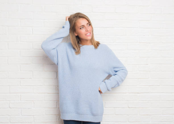 Beautiful young woman over white brick wall confuse and wonder about question. Uncertain with doubt, thinking with hand on head. Pensive concept.