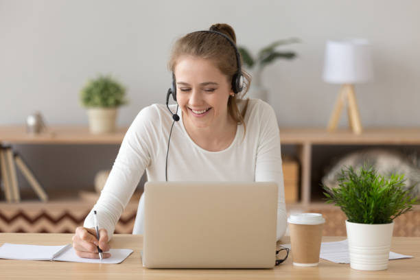 sonriente mujer vistiendo auricular escritura notas estudiando en línea en portátil - help palabra en inglés fotografías e imágenes de stock