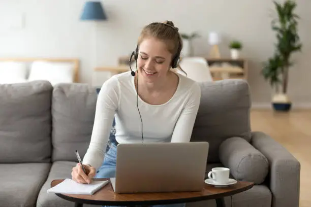 Happy girl interpreter in headset translate write notes listen audio course study work on laptop, smiling teen student wearing headphones learning online training class, distance education concept