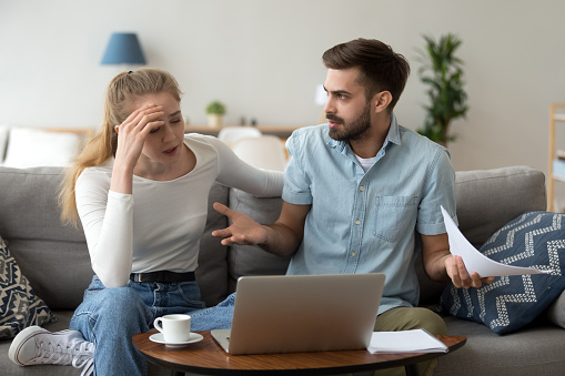 Stressed unhappy couple arguing about huge expenses with laptop and papers, angry husband blaming wife of overspending debt, family having conflict fight about wasting money financial problem at home