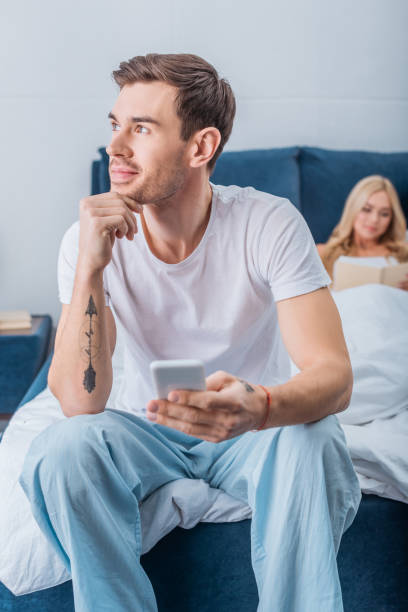 istock pensive man sitting with hand on chin and using smartphone while girlfriend reading book in bed behind 1124672547
