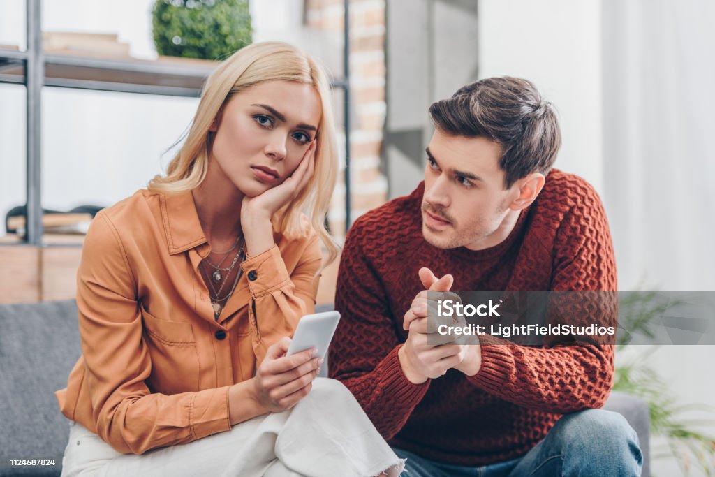 husband looking at unhappy young woman holding smartphone and looking at camera at home, relationship problem concept Envy Stock Photo