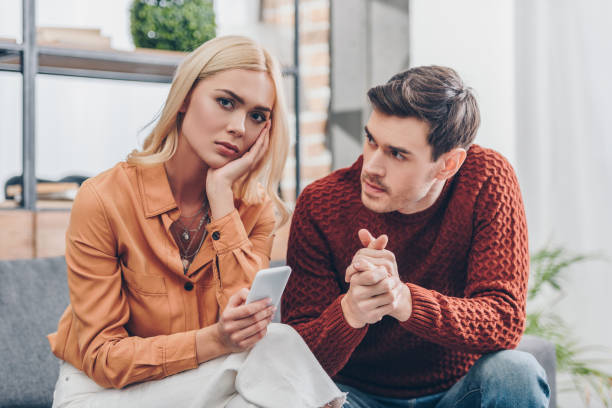 Husband Looking At Unhappy Young Woman Holding Smartphone And Looking At Camera At Home Relationship Problem Concept Stock Photo - Download Image Now - iStock