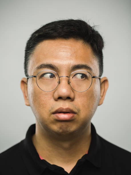 Close up portrait of asian man with blank expression looking to the side against white gray background. Vertical shot of chinese real people observing in studio with black hair and glasses. Photography from a DSLR camera. Sharp focus on eyes.