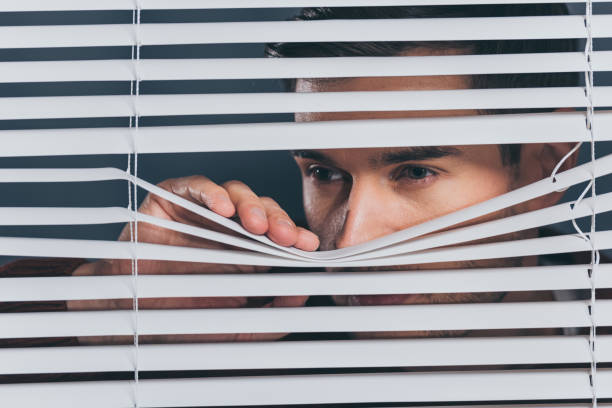 istock suspicious young man looking away and peeking through blinds 1124692184