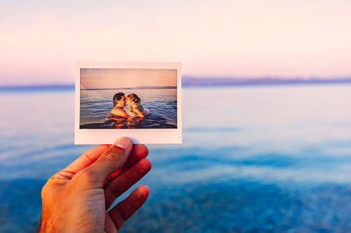 Father is connecting his polaroid photograph with the present scene. Picture of his daughter and himself enjoying together at the sea. Authentic moment, real people.