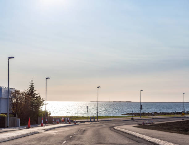Empty curve road near sea. Asphalt highway with street lights against sky. Traffic cones can be seen on roadside.