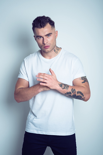 Studio portrait of angry tattooed young man wearing white t-shirt standing against grey background.