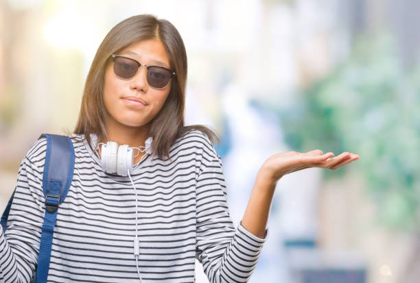 young asian student woman wearing headphones and backpack over isolated background clueless and confused expression with arms and hands raised. doubt concept. - head and shoulders audio imagens e fotografias de stock