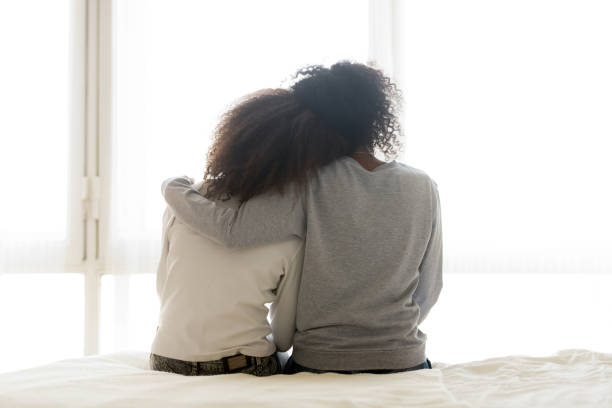 Back view of loving mom hug teen daughter Back view of loving African American mother hug teen daughter sitting on bed, caring black mom embrace child, relaxing in bedroom looking in window, parent comfort teenager caressing at home life events photos stock pictures, royalty-free photos & images
