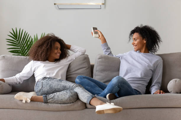 Happy mom and daughter relaxing on couch under air conditioner Smiling African American mom and daughter sit on cozy couch relaxing, operating air conditioner with remote controller, happy teenager and mother or nanny rest on sofa at home, enjoying fresh air air conditioner family stock pictures, royalty-free photos & images