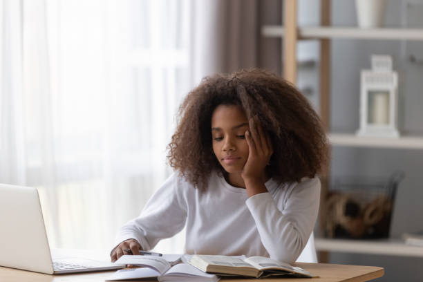 Dreamy black teenager distracted from preparing homework Dreamy African American teenage girl sit at table with laptop and textbooks, distracted from doing homework, black teenager dream or think when preparing housework with handbook and notebook wasting time stock pictures, royalty-free photos & images