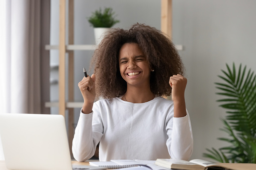Excited African American teenage girl gesture yes finishing homework, preparing project or exam with textbook, happy black teen pupil satisfied with grade or achievement, doing school task at desk