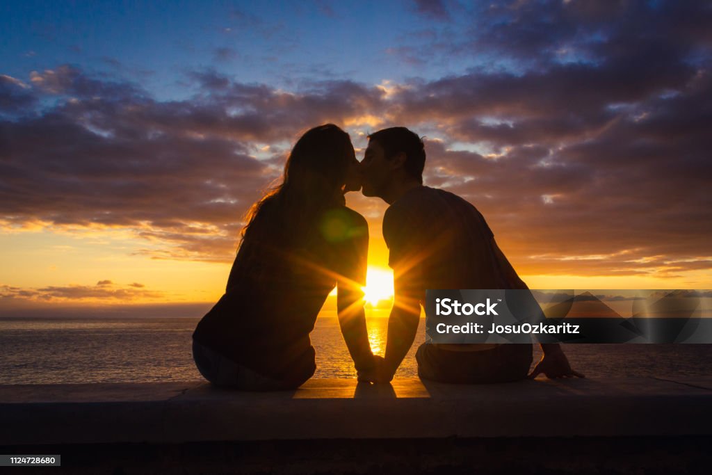Man and woman sitting by the sea kissing at sunset at Meloneras beach walk, Gran Canaria Couple silhouette enjoying romantic colorful twilight. Valentines Day, honeymoon romantic date concepts Couple - Relationship Stock Photo