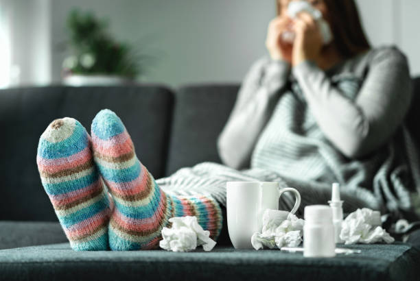 kranke frau mit grippe, kälte, fieber und husten auf couch zu hause sitzen. kranke person nase und niesen mit gewebe und taschentuch. wollsocken und medizin. infektion im winter. - krankheit stock-fotos und bilder