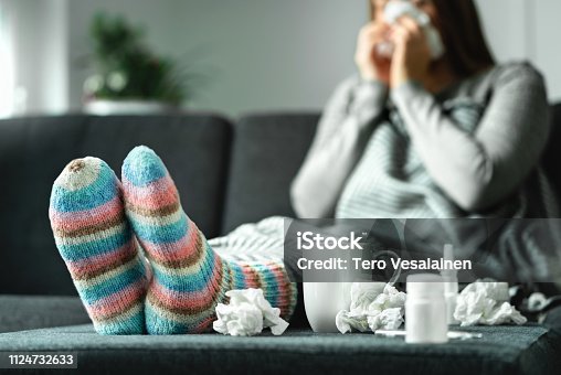 istock Sick woman with flu, cold, fever and cough sitting on couch at home. Ill person blowing nose and sneezing with tissue and handkerchief. Woolen socks and medicine. Infection in winter. 1124732633