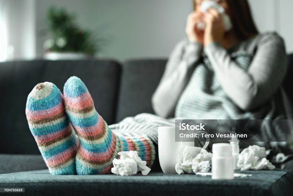 Sick woman with flu, cold, fever and cough sitting on couch at home. Ill person blowing nose and sneezing with tissue and handkerchief. Woolen socks and medicine. Infection in winter. Sick woman with flu, cold, fever and cough sitting on couch at home. Ill person blowing nose and sneezing with tissue and handkerchief. Woolen socks and medicine. Infection in winter. Resting on sofa. Cold And Flu Stock Photo
