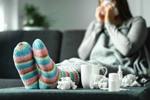 Sick woman with flu, cold, fever and cough sitting on couch at home. Ill person blowing nose and sneezing with tissue and handkerchief. Woolen socks and medicine. Infection in winter. stock photo