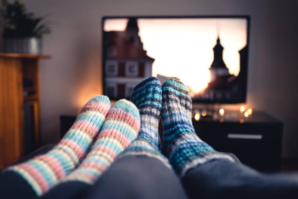 Photo of Couple with socks and woolen stockings watching movies or series on tv in winter. Woman and man sitting or lying together on sofa couch in home living room using online streaming service.