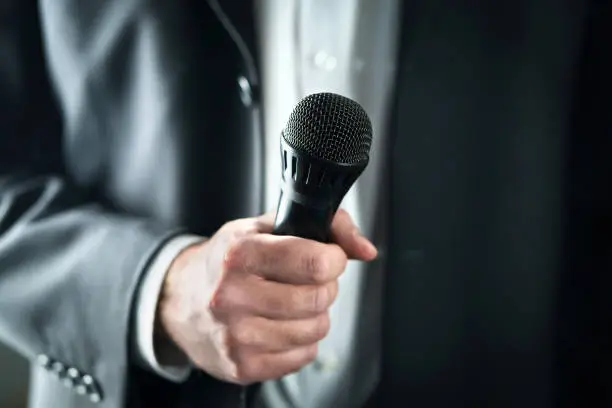 Business man holding microphone. Public speaking and giving speech in suit for audience concept. Fiance, host or best man giving toast with mic in wedding. Karaoke, talent show or singing contest.