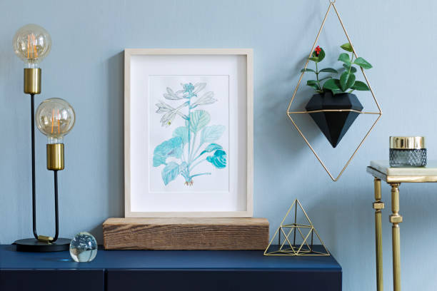 istock Home interior with poster mock up with wooden frame, table lamp, gold pyramid, table, accessories and hanging plant in geometric pot on the grey wall background. Minimal concept of navy blue shelf. 1124734337
