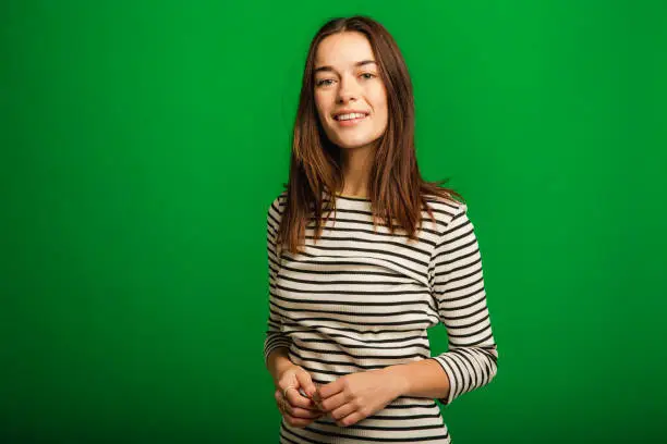 Portrait of a young woman looking at the camera while clasping her hands. She is standing in front of a green studio background.
