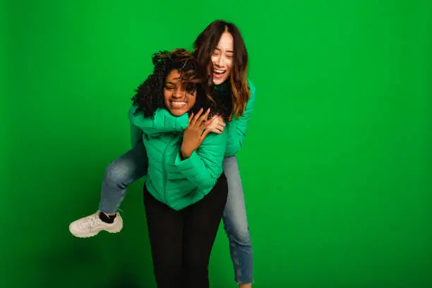 Two female friends having fun infront of a green background in a studio.