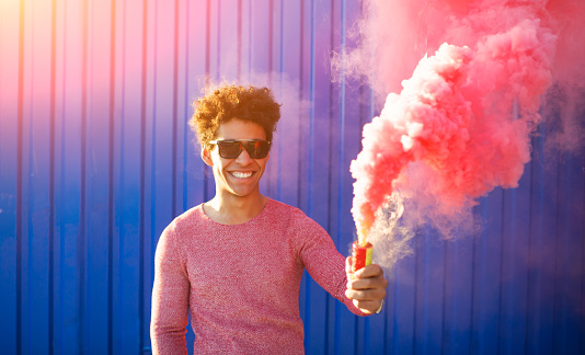 Colorful young black hipster man smiling and holding a  pink smoke bomb over a blue color background. Youth and lifestyle concept