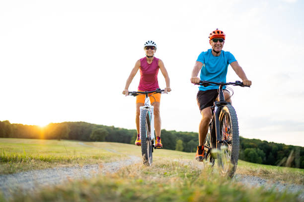 40-50 years old sporty couple cycling on electric mountain bikes in rural environment low angle view athletic sporty fit happy smiling  woman and man couple friends in their fourties cycling with their electric mountain bicycles on gravel path through grassland rural landscape on sunny summer afternoon sunset athleticism photos stock pictures, royalty-free photos & images