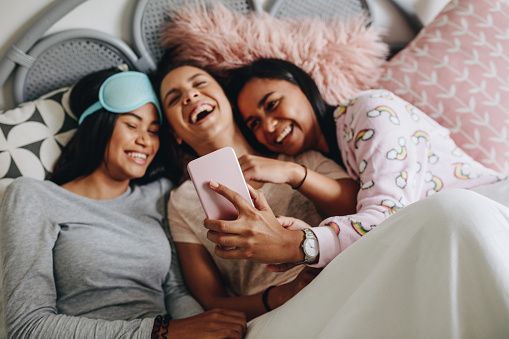 Young girls lying on bed during a sleepover laughing while looking at mobile phone. Girl taking selfie using mobile phone lying on bed with friends at a sleepover.