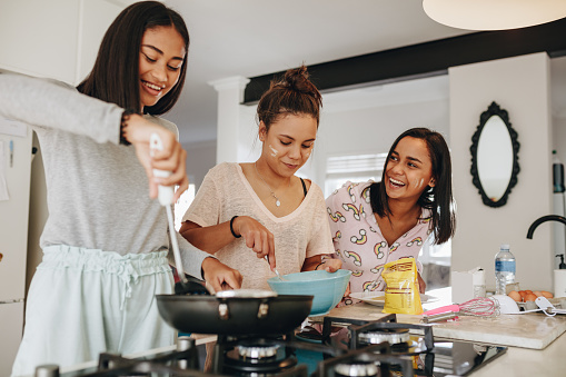 Young girls preparing breakfast in kitchen. Smiling girl cooking food while her friends help her in kitchen.