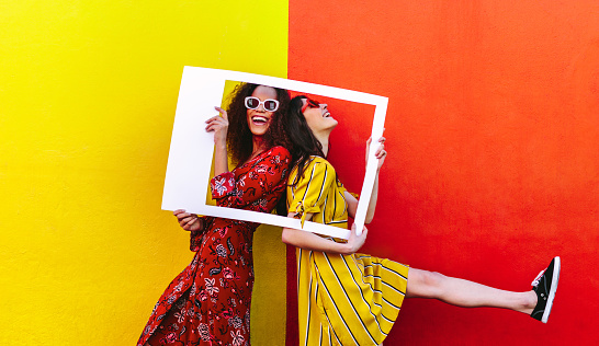 Smiling women with empty photo frame against colored wall. Cheerful girls wearing colorful dresses standing with a picture frame.