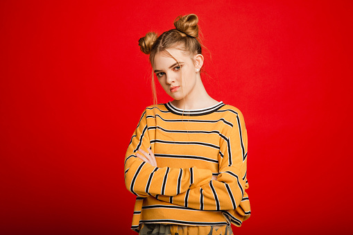 Teenage girl with her arms crossed and a serious facial expression while looking at the camera. She is standing infront of a red studio background.