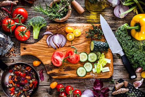 Top view of a rustic wooden table filled with fresh organic vegetables ready for cooking. At the center of the frame is a wooden cutting board filled with some sliced vegetables and a kitchen knife. The composition includes garlic, bell pepper,  tomatoes,  cucumber, broccoli, onion, lettuce, and chili peppers. An olive oil bottle and a mortar are at the top of the frame. DSRL studio photo taken with Canon EOS 5D Mk II and Canon EF 100mm f/2.8L Macro IS USM