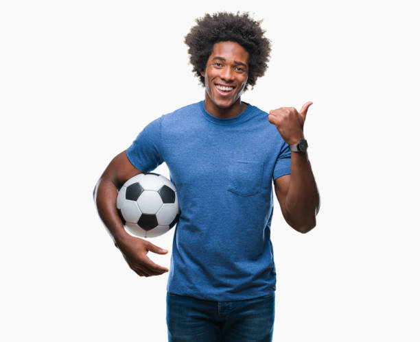 afro americano homem segurando uma bola de futebol sobre fundo isolado apontando e mostrando com o polegar até o lado com uma cara feliz sorrindo - football human hand holding american football - fotografias e filmes do acervo