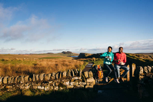 Two young adults sitting on a fence in the Northumberland, UK countryside, eating lunch.