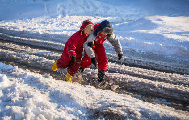 Two boys in rainboots playing with toy boat an the creek of melting snow at sunny spring day, outdoors