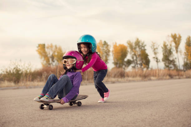 Two Girls Racing on a Skateboard Two young girls are racing on a skateboard. One girl is pushing the other and they are going really fast on a rural road. They are young adventurers wearing helmets and flight goggles and are ready to beat all the boys in the race. Image taken in Utah, USA. pushing stock pictures, royalty-free photos & images