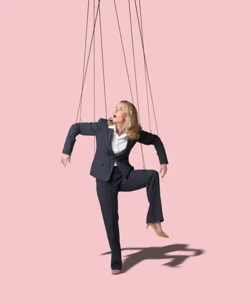 A businesswoman looks up as she is controlled by the strings of a marionette against a pink background.