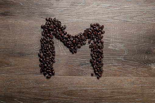 Dark roasted coffee bean arranged on a wooden table in the shape of text alphabet letter M