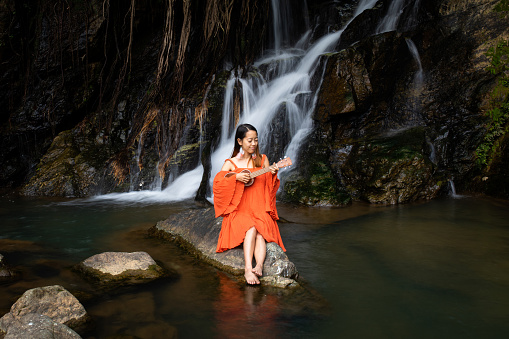 A young pretty Japanese lady playing Ukelele by a waterfall