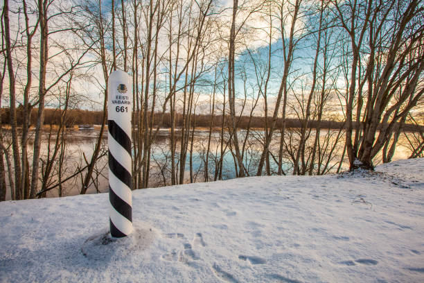 Estonian border marker stands in snow near a river, with bare trees and a colorful winter sky in the background.
