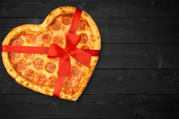 Pizza pepperoni shaped heart with red ribbon bow for Valentine's Day on rustic dark black background. Top View. Flat lay
