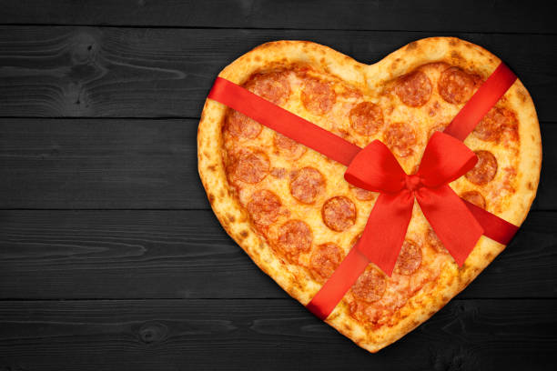 istock Pizza pepperoni shaped heart with red ribbon bow for Valentine's Day on rustic dark black background. Top View. Flat lay 1124998888
