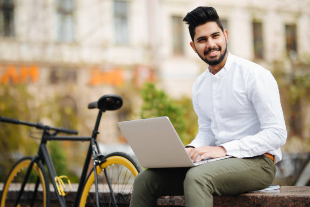 istock Confident young stylish indian man working on laptop computer while sitting outdoors with bicycle 1125004787