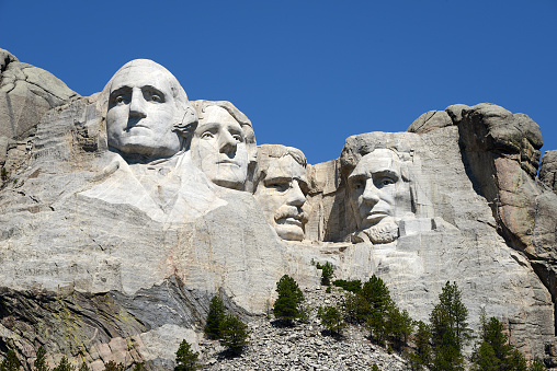 Front view of Mount Rushmore. The carved faces of the four historical figures, George Washington, Thomas Jefferson, Theodore Roosevelt and Abraham Lincoln are framed by a brilliant stormy sky and tree branches. Bright green coniferous trees in the foreground provide contrast to the granite stone.