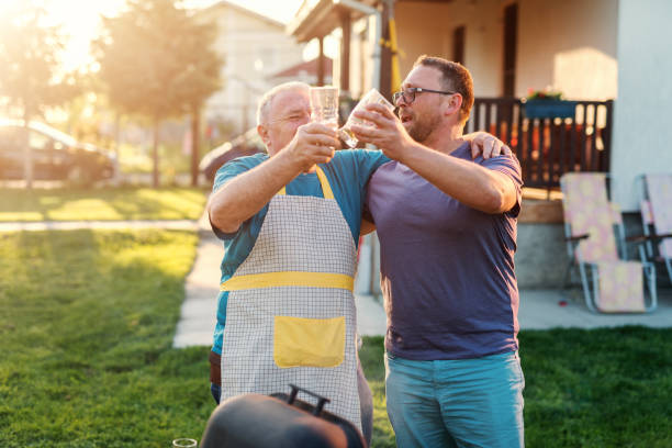 Father in law and his son in law making a toast with beer and hugging while standing next to grill. Family gathering concept. Father in law and his son in law making a toast with beer and hugging while standing next to grill. Family gathering concept. father in law stock pictures, royalty-free photos & images