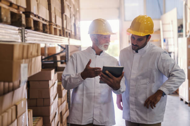 istock Two coworkers in white uniform and with yellow helmets on heads looking at tablet while standing in storage. Quality control concept. 1125026032