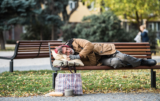 A homeless beggar man with a bag lying on bench outdoors in city, sleeping. Copy space.